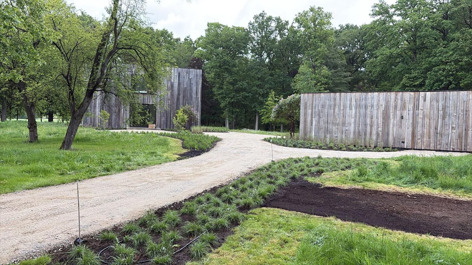 Grindoprit tussen nieuw aangeplante borders naar een woning en bijgebouw met houten gevels aan de bosrand